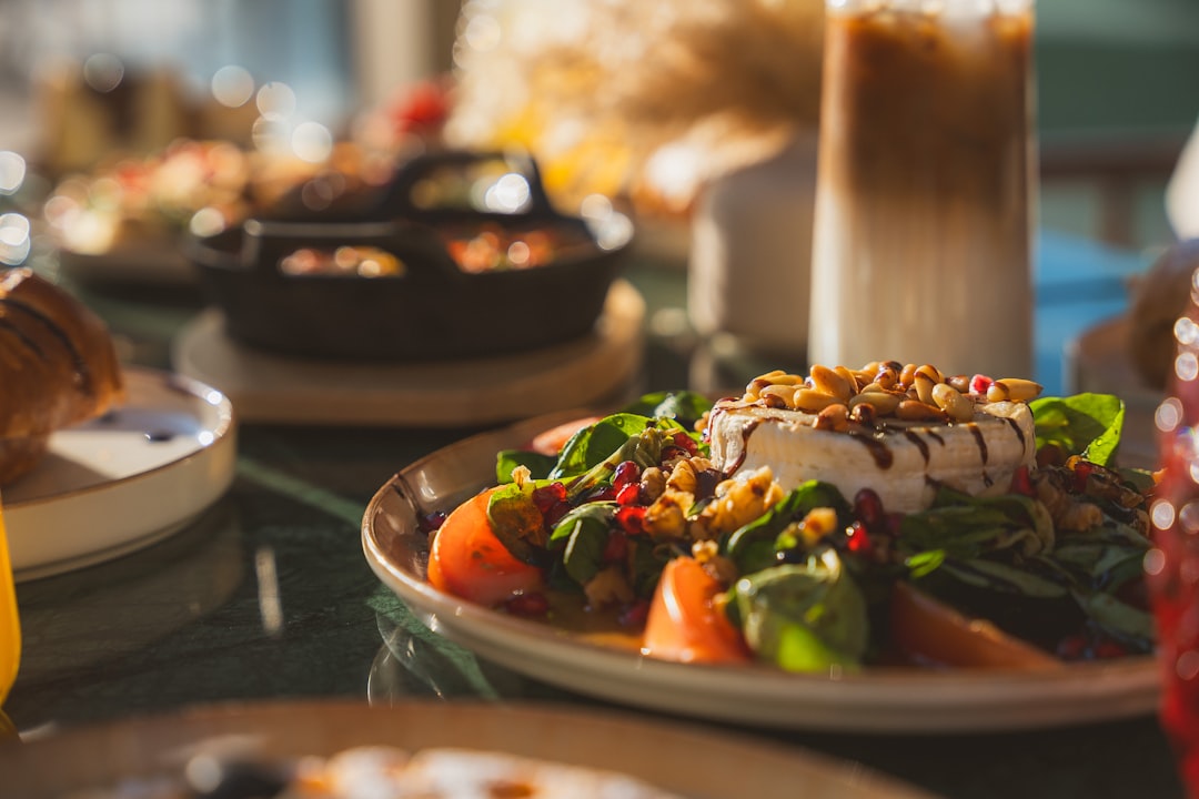 A close-up of a fresh salad topped with cheese, nuts, and pomegranate seeds on a dining table with blurred food and drinks in the background.