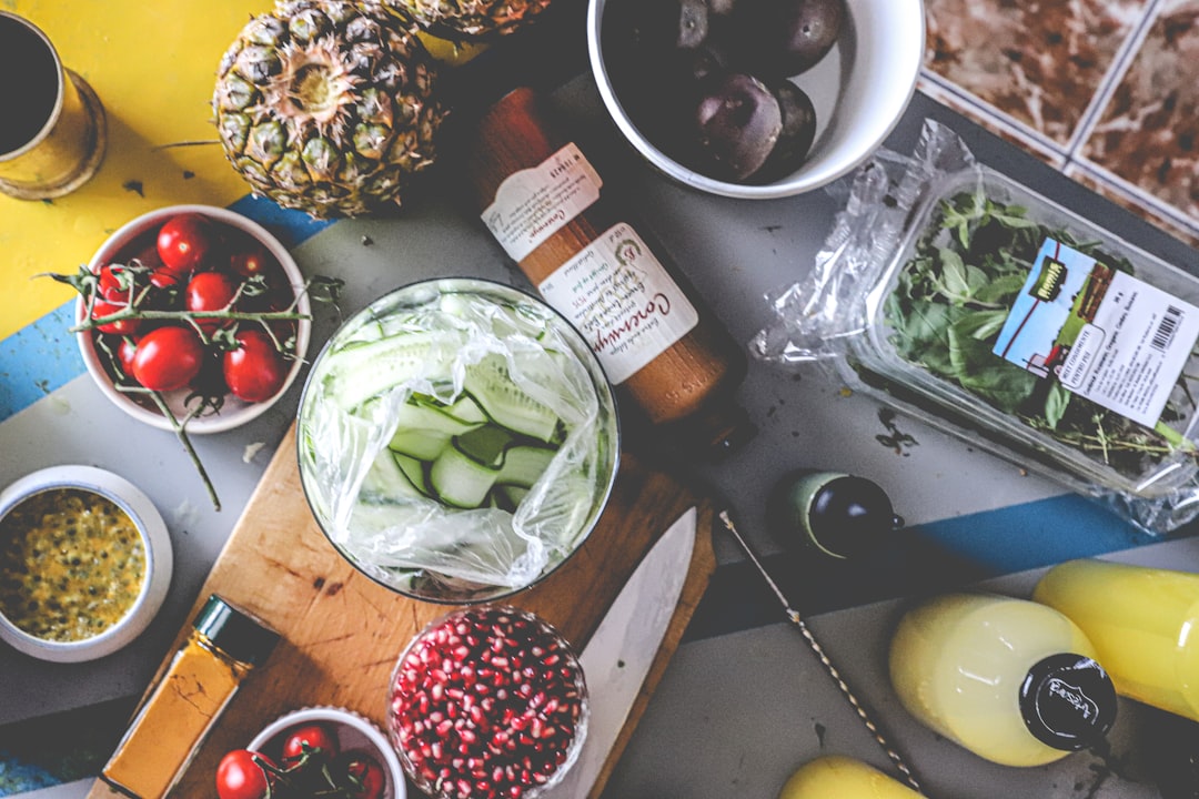 A top-down view of various fresh food and drink ingredients, including pineapple, tomatoes, cucumber, pomegranate seeds, and basil, laid out on a table.