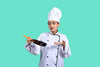 "Female chef in a white uniform and hat holding a frying pan against a turquoise background."