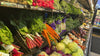 **Alt text:** Fresh produce display in a grocery store with colorful vegetables including carrots, radishes, cabbage, broccoli, and leafy greens neatly arranged on shelves.